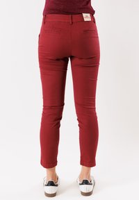 Pantaloni rossi alla caviglia realizzati in tessuto elasticizzato, con tasche posteriori e un'etichetta con il brand in vita, abbinati a sneaker bianche e dettagli neri.
