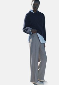 Pulover tricotat în nuanța bleumarin, cu manșete cu nervuri, purtat peste o cămașă albă și pantaloni bej largi. Textura netedă și designul cu picioare largi accentuează stilul.