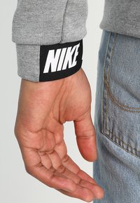 Grå långärmad skjorta med ribbstickade muddar; svart etikett på ärmen med vit "NIKE"-logotyp. Hand placerad nära en ljusblå denimbakgrund.
