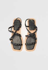 Sandales plates noires avec des brides entrecroisées, ornées de clous et d'une semelle beige contrastante. Matériau en cuir lisse avec une forme rectangulaire.