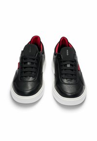 Mustat nahkakengät, joissa on teksturoitu pinta, mustat nauhat, punaiset yksityiskohdat ja valkoinen kumipohja. Logo kielessä ja sivussa.