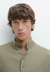 Beige strik polo skjorte med høj krave, prydet med tre grå knapper og et struktureret stof. Modellen har kort, bølget hår og et neutralt udtryk.