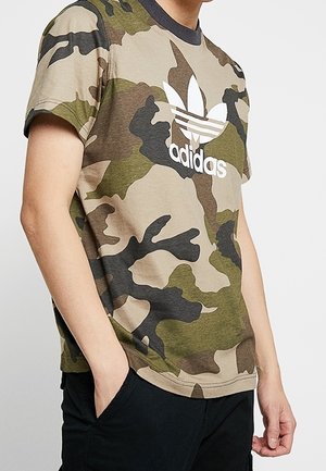 Person trägt ein grünes, braunes und schwarzes Camouflage Adidas T-Shirt mit weißem Logo, Hände in schwarzen Hosentaschen, steht vor schlichtem Hintergrund.