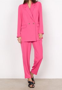 Jasnoróżowy żakiet o podwójnym zapięciu, złote guziki oraz dwie przednie kieszenie, w połączeniu z pasującymi różowymi spodniami, gładka tekstura i luźny fason.