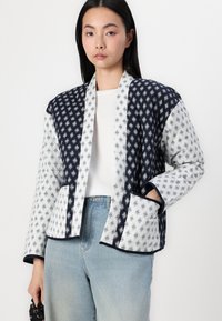 Quilted jacket in navy and white, featuring a geometric pattern, open front, two pockets, and a relaxed fit with contrasting trim.