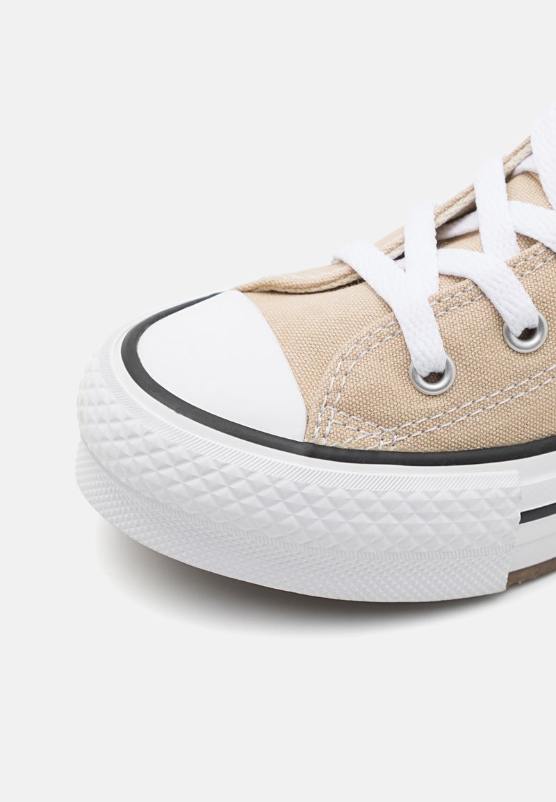 Zapatilla de lona beige con suela de goma blanca, ribete negro y punta redonda. Cuenta con cordones blancos y ojales de metal para el cierre.