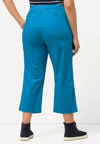 Teal geknipte broeken met een elastische tailleband, een gladde textuur en zijsplitten aan de zoom. Gepaard met zwarte hoge sneakers.