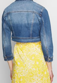 Skuren blå denimjacka med långa ärmar och detaljer på manschetterna, ihop med en gul blommönstrad kjol.