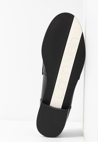 Semelle d'une chaussure habillée noire avec une bande blanche comportant le logo Calvin Klein embossé, présentée sur un fond blanc uni.