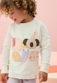 Krämfärgad långärmad skjorta med färgglada djurapplikationer: en koala, snigel och katt, med texturerade tyger och lekfulla mönster.