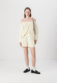 Ungdommelig kvinne iført off-shoulder løs kremfarget topp og matchende shorts, svarte loafers, hvite sokker, med en brun skulderveske.