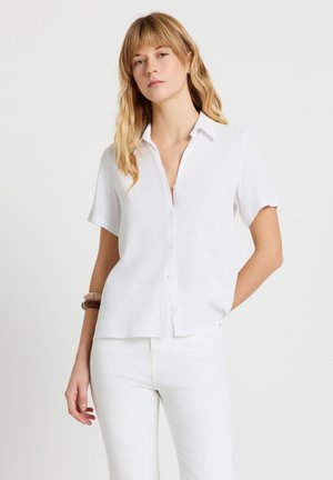 Frau trägt ein weißes kurzärmeliges Hemd mit Knöpfen und weiße Hose, eine Hand in der Tasche, neutraler Gesichtsausdruck.