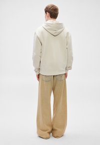 Világos bézs kapucnis pulóver középső varrással, nagy kapucnival és bordázott mandzsettákkal, laza, világosbarna, bő szárú nadrággal, amelyen hátsó zsebek találhatók.