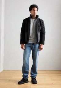 Giacca in pile nera con accenti grigi, felpa grigia, jeans blu e scarpe nere. Design elegante, tasche con zip e tessuto testurizzato.