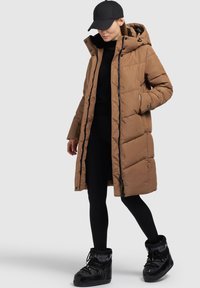 Braune Daunenjacke mit Reißverschluss, Kapuze und Seitentaschen. Glänzende schwarze Schneestiefel und schwarze Leggings runden das Outfit ab.