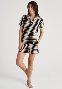 Gestreept katoenen polo- en shortset in zwart en crème. Kenmerkt zich door een kraag. Materiaal heeft een zachte textuur. Model poseert blootvoets.
