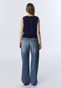 Person with long hair wearing a sleeveless navy sweater and loose-fitting faded blue jeans, standing facing away against a white background.