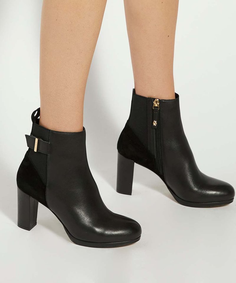 Bottines en cuir noir avec talons blocs, fermetures éclair latérales et lanières décoratives à l'arrière, portées aux pieds sur un fond uni.