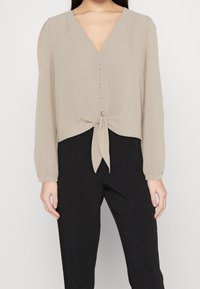 Donna che indossa una camicia beige a maniche lunghe con bottoni frontali e un dettaglio a fiocco, abbinata a pantaloni neri a vita alta su uno sfondo neutro.