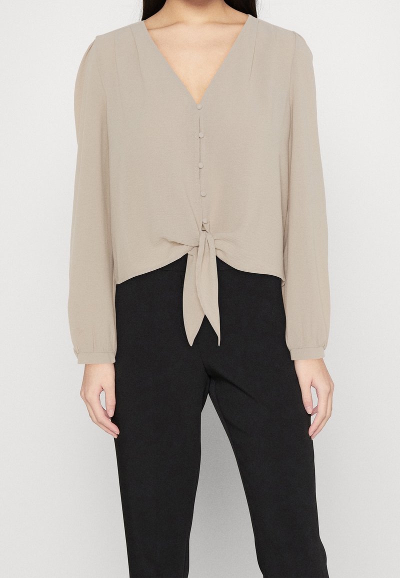 Donna che indossa una camicia beige a maniche lunghe con bottoni frontali e un dettaglio a fiocco, abbinata a pantaloni neri a vita alta su uno sfondo neutro.