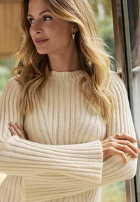 Pull en maille côtelée crème avec un col haut et des manches larges. Accents de couture délicate et texture subtile. Boucles d'oreilles en or visibles.