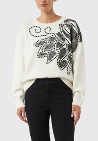 Biały dziany sweter z czarnym abstrakcyjnym kwiatowym wzorem zdobionym koralikami, okrągłym dekoltem i luźnym krojem, zestawiony z czarnymi dżinsami.