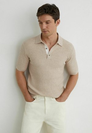 Homme portant un polo beige à manches courtes texturé et un pantalon crème, debout les mains dans les poches contre un mur blanc.