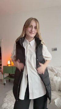 Schwarze Steppweste über einem weißen Hemd mit Knöpfen, das einen hohen Kragen, eine glatte Textur und Druckknopfverschlüsse aufweist. Schwarze Hosen runden das Outfit ab.