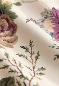 Tessuto floreale con uno sfondo crema, fiori viola e azzurri, accenti verdi fogliari e una texture satinata liscia.
