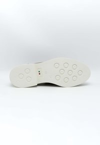 Semelle de chaussure marron avec une base en caoutchouc blanc, présentant des motifs de grip circulaires et une petite étiquette verte, blanche et rouge près du centre.