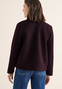 Donna che indossa una giacca in pile testurizzata di un viola scuro con maniche lunghe e jeans blu, vista da dietro su uno sfondo neutro.