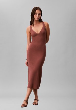 Vrouw die tegen een effen achtergrond staat, gekleed in een mouwloze, roestkleurige, geribbelde midi-jurk en zwarte sandalen met open tenen.