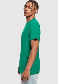 Camiseta de algodón verde con mangas cortas, cuello redondo y detalle de logo en el frente. Forma simple y ajustada con una textura suave.