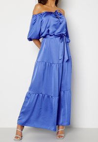 Robe maxi en satin bleu, décolletée à épaules nues, avec manches courtes bouffantes, encolure nouée et ourlet à volants. Comprend une ceinture assortie à la taille.