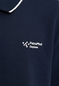 Námořnická modrá polo košile s texturovanou tkaninou, s bílým límečkem a potištěným logem, které obsahuje text "Petrol Ind Daytona."