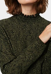 Grönmönstrad topp med en volangkrage och långa ärmar, tillverkad av ett mjukt strukturerat tyg med ett zigzag-mönster.