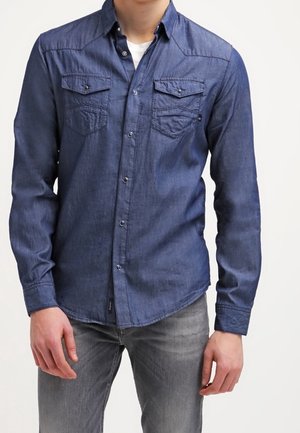 Sininen farkkupaita napilla, jossa on pitkät hihat, kaksi rintataskua, terävä kaulus ja sileä pinta. Yhdistetty harmaisiin farkkuihin.