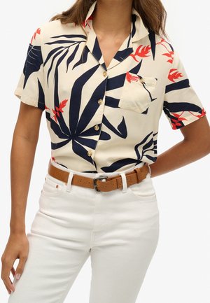 Kobieta ubrana w kremową koszulę z krótkim rękawem ozdobioną granatowo-czerwonym tropikalnym liściastym wzorem, wpuszczoną w białe spodnie z beżowym skórzanym paskiem.