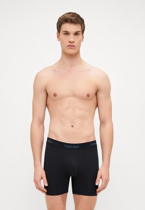 Čierne bavlnené boxerky s logom na páse, ktoré majú priliehavý strih a hladký povrch. Model stojí na jednoduchom bielom pozadí.