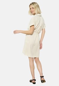 Beige raidallinen mekko, jossa on lyhyet hihat ja vyötärölle sidottava nauha; siinä on kaulus ja hieman avara helma. Mustat kengät, joissa on satiinirusetit.