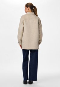 Beige ulljacka med krage, långa ärmar och knappar vid ärmsluten, kombinerad med vidbeniga mörka jeans i denim och svarta skor.