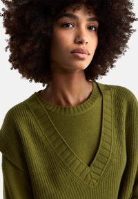Zelený pletený svetr s hlubokým výstřihem do V, zdobený žebrovanými detaily a strukturovanou látkou. Rukávy jsou volné.