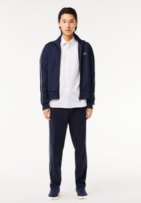 Trening bleumarin cu jachetă cu zip și pantaloni, cu dungi albe pe laterale. Jacheta are guler ribat și un detaliu cu logo. Sub jachetă se află o cămașă albă cu nasturi.