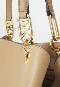 Sac à main en cuir beige avec quincaillerie dorée, finition texturée et clips métalliques décoratifs. Le sac a une forme arrondie et des sangles robustes.