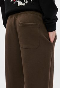 Καφέ φόρμες με υφή, διαθέτοντας λοξό πίσω τσέπη και ελαστική μέση, συνδυασμένες με μαύρο μπλουζάκι με μακριά μανίκια.