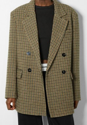 Persoon draagt een dubbelgeknoopt bruin en beige houndstooth colbert over een zwarte cropped top en zwarte broek met een witte riem.