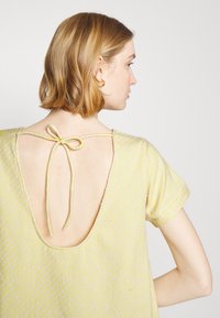 Camisola amarela com padrão e mangas curtas. Apresenta um decote em V profundo nas costas com um fecho de laço. Tecido texturizado com pontos subtis em tons mais claros.
