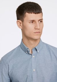 Camisa abotoada azul claro feita de um tecido com uma textura suave. Apresenta um colarinho pontiagudo e botões brancos ao longo da pala.