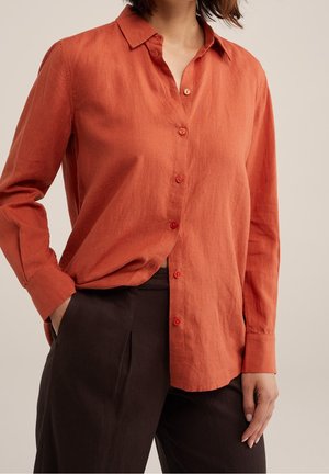 Femme portant une chemise orange brûlée à manches longues boutonnée et rentrée dans un pantalon noir taille haute, avec une main dans la poche.