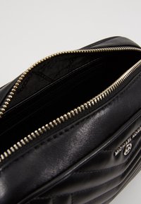 Schwarze Ledertasche mit gestepptem Muster, silbernem Reißverschluss und Innenfächern. Branding auf der Seite sichtbar. Schlankes, strukturiertes Design.